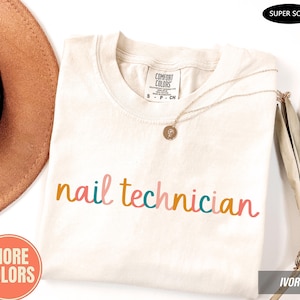 May include: An ivory short-sleeved t-shirt with the text "nail technician" printed in a multicoloured cursive font. The shirt features a Comfort Colors brand label at the neck, a thin gold-toned necklace, and a black "SUPER SOFT" tag in the corner.
