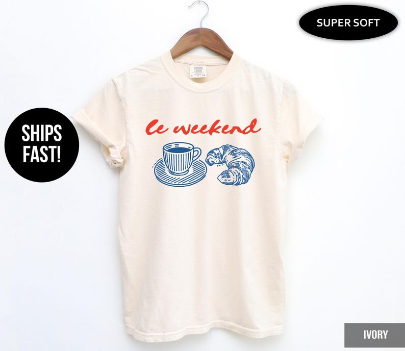 May include: Ivory-colored t-shirt with the words "Le weekend" in red script. Below the text is a blue and white illustration of a coffee cup on a saucer and a croissant. The shirt is hanging on a wooden hanger.