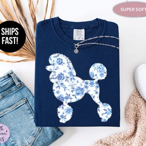 May include: A navy blue sweatshirt featuring a white poodle silhouette filled with a blue floral pattern. A silver necklace with a charm rests on the shirt. The words "SUPER SOFT" are in a pink oval. The words "SHIPS FAST!" are in a black circle.