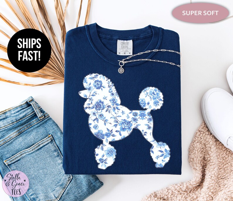 May include: A navy blue sweatshirt featuring a white poodle silhouette filled with a blue floral pattern. A silver necklace with a charm rests on the shirt. The words "SUPER SOFT" are in a pink oval. The words "SHIPS FAST!" are in a black circle.