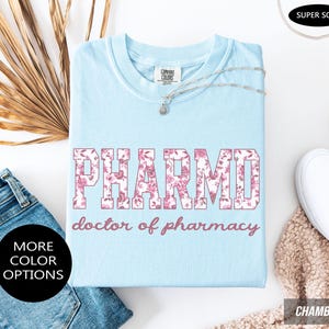 May include: A light blue t-shirt featuring the text PHARMD in a pink floral pattern, with the words doctor of pharmacy printed below in a matching pink script font. The shirt has a crew neckline and a Comfort Colors brand label at the neck.