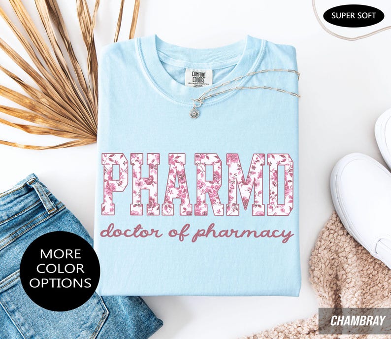 May include: A light blue t-shirt featuring the text PHARMD in a pink floral pattern, with the words doctor of pharmacy printed below in a matching pink script font. The shirt has a crew neckline and a Comfort Colors brand label at the neck.
