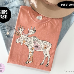 May include: A terracotta-colored short-sleeve t-shirt featuring a large graphic of a moose silhouette filled with a detailed floral pattern in shades of pink, orange, and green. The shirt has a crew neckline and a Comfort Colors brand label.