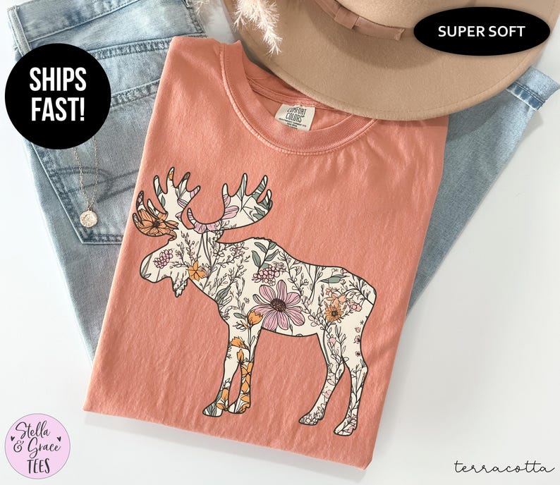 May include: A terracotta-colored short-sleeve t-shirt featuring a large graphic of a moose silhouette filled with a detailed floral pattern in shades of pink, orange, and green. The shirt has a crew neckline and a Comfort Colors brand label.