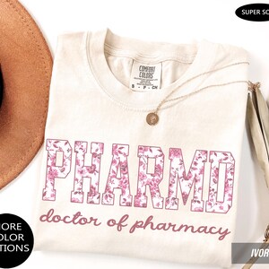 May include: An ivory t-shirt featuring the text PHARMD in a block font filled with a pink floral pattern, with the text doctor of pharmacy written below in a cursive script. The shirt has a Comfort Colors label and a round neckline.
