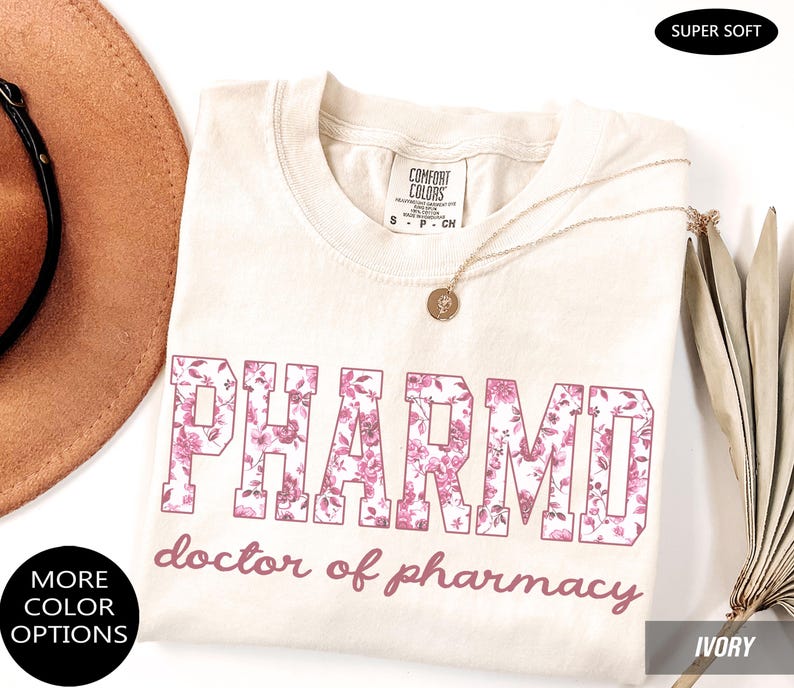 May include: An ivory t-shirt featuring the text PHARMD in a block font filled with a pink floral pattern, with the text doctor of pharmacy written below in a cursive script. The shirt has a Comfort Colors label and a round neckline.