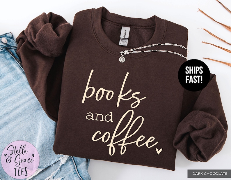 May include: Dark chocolate-colored sweatshirt with the words "books and coffee" in a cream-colored script. A silver necklace with a pendant is draped on the sweatshirt. The sweatshirt is folded next to a pair of blue jeans.