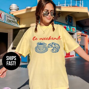 May include: A light yellow t-shirt with the text "le weekend" in red and a blue graphic of a coffee cup and croissant. The shirt has a relaxed fit and is labeled "Super Soft".