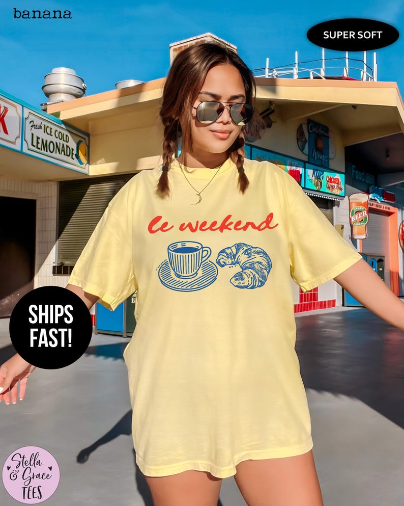 May include: A light yellow t-shirt with the text "le weekend" in red and a blue graphic of a coffee cup and croissant. The shirt has a relaxed fit and is labeled "Super Soft".