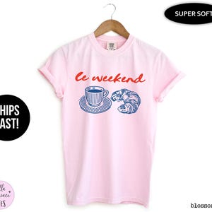 May include: A light pink t-shirt with the words "le weekend" in red script. The shirt features a blue and white illustration of a coffee cup and a croissant. The shirt is hanging on a wooden hanger.