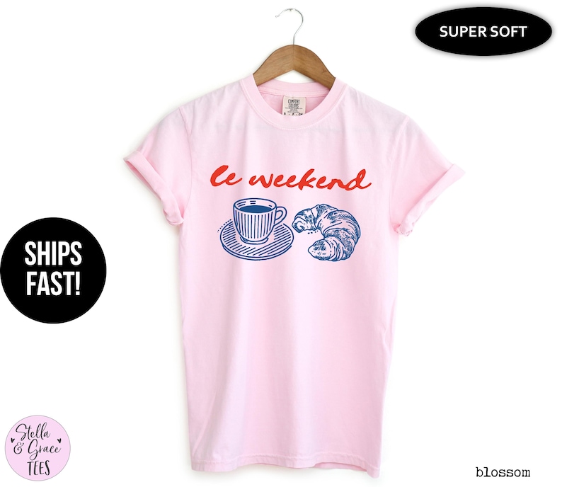 May include: A light pink t-shirt with the words "le weekend" in red script. The shirt features a blue and white illustration of a coffee cup and a croissant. The shirt is hanging on a wooden hanger.