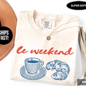 May include: Ivory-colored t-shirt with the words "Le weekend" in red script. Below the text is a blue and white illustration of a coffee cup and a croissant. The shirt is labeled "Super Soft."