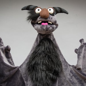 May include: A gray puppet with a furry chest and large hands. The puppet has a brown nose, large white eyes, and a pink tongue. It has black horns and a black furry patch on its head. The puppet is set against a gray background.