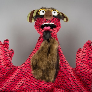 May include: A hand puppet featuring a bright red textured body with black markings, a brown faux fur chest patch, and a black goatee. The puppet has large white eyes with yellow pupils, a wide open mouth with white teeth, and striped horns.