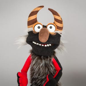 May include: A hand puppet featuring a black face with a large brown nose, white eyes, and a row of white teeth. It has two curved horns with brown and tan stripes, grey faux fur hair on the sides, and a body covered in dark grey fur with red arms.