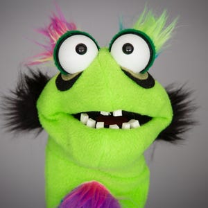 May include: A bright lime green puppet featuring large white eyes with black pupils, black fur accents on the sides, and a tuft of multicolored rainbow hair on top. The puppet has a wide open mouth revealing small white square teeth.