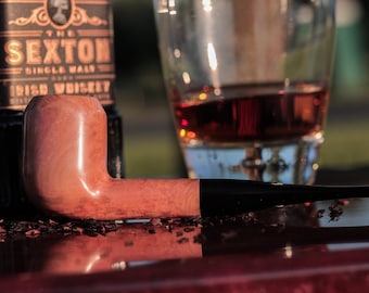 Op de afbeelding: Een houten tabakspijp met een bruine kom en een zwarte steel. De pijp ligt op een donker oppervlak met wat losse tabak eromheen. Op de achtergrond staat een glas met een amberkleurige vloeistof.