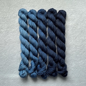 May include: Five skeins of yarn arranged in a vertical row, displaying a gradient of blue colors. The sequence transitions from a light denim blue on the left to a deep, dark navy blue on the right, with each skein featuring a twisted texture.
