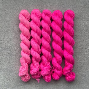 Könnte beinhalten: Fünf Stränge leuchtend rosa Garn, vertikal angeordnet. Die Garnfarben variieren von hellrosa bis tiefrosa und erzeugen einen Farbverlauf. Das Garn ist eng gewickelt, wodurch die Textur der Fasern sichtbar wird. Ideal für Strick- oder Häkelprojekte.