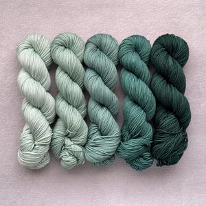 Könnte beinhalten: Fünf Stränge Garn in verschiedenen Grüntönen, von hellem Mint bis tiefem Waldgrün, liegen auf einer hellvioletten Oberfläche. Das Garn ist eng gewickelt und zeigt die Textur- und Farbvariationen, ideal für Strick- oder Häkelprojekte.