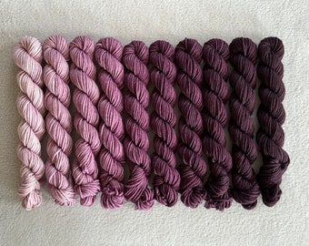 May include: A set of ten skeins of yarn in shades of pink and purple, ranging from light to dark. The yarn is arranged in a gradient, with the lightest shade at the top and the darkest shade at the bottom.