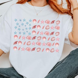 May include: White t-shirt featuring a design of a flag made of blue stars, pink seashells, and pink starfish. The shirt is a casual, comfortable style, perfect for a beach or summer theme.