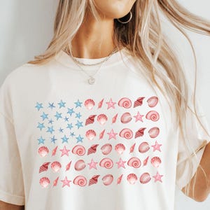 May include: Cream-colored t-shirt featuring a design resembling the American flag, with blue stars and red seashells and starfish. The design is a unique take on patriotic themes, perfect for beach lovers.