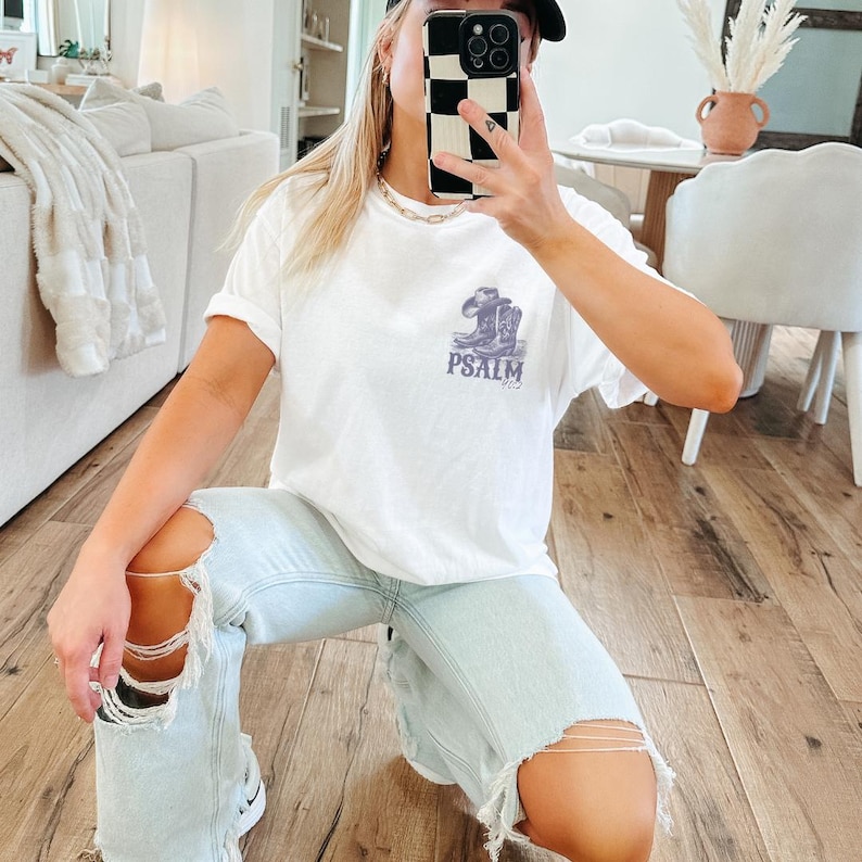 K&ouml;nnte beinhalten: Ein wei&szlig;es T-Shirt mit einem Aufdruck von Cowboystiefeln und dem Text "PSALM 23". Die Person, die das Shirt tr&auml;gt, tr&auml;gt auch hellblaue Jeans mit Destroyed-Details.