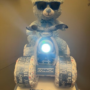 May include: A blue teddy bear wearing sunglasses is riding a four-wheeled diaper cake. The cake is decorated with blue and white ribbons and the words "Baby Joy It's a Boy". The cake is labeled "CONNOR".
