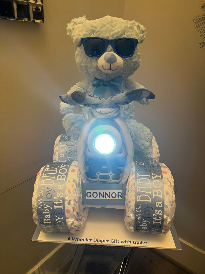 May include: A blue teddy bear wearing sunglasses is riding a four-wheeled diaper cake. The cake is decorated with blue and white ribbons and the words "Baby Joy It's a Boy". The cake is labeled "CONNOR".