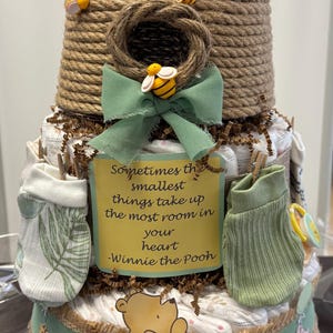 May include: A three-tiered baby shower diaper cake, resembling a beehive, decorated with bee accents and a Winnie the Pooh theme. Includes a green bow, baby clothes, and a sign with a quote from Winnie the Pooh.