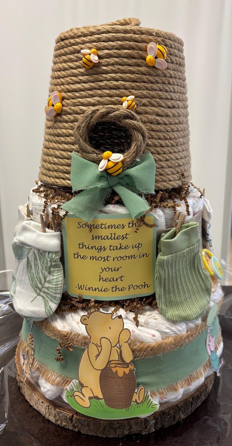 May include: A three-tiered baby shower diaper cake, resembling a beehive, decorated with bee accents and a Winnie the Pooh theme. Includes a green bow, baby clothes, and a sign with a quote from Winnie the Pooh.