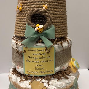 May include: A three-tiered baby shower cake decorated with a beehive theme. The cake features a Winnie the Pooh theme, with a quote on a yellow tag. Decorative bees and a pacifier are included.