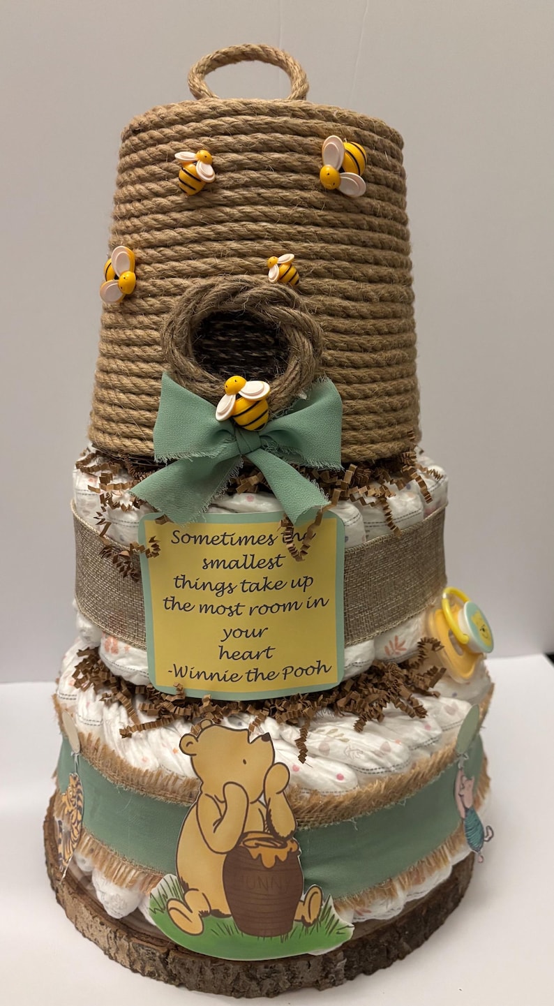 May include: A three-tiered baby shower cake decorated with a beehive theme. The cake features a Winnie the Pooh theme, with a quote on a yellow tag. Decorative bees and a pacifier are included.