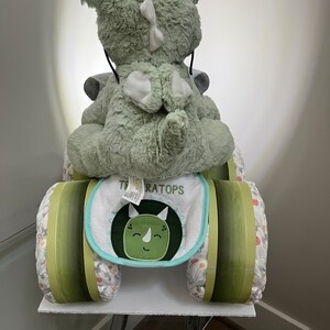 May include: A green plush dinosaur toy riding a diaper cake decorated with green and white stripes. The cake is labeled "TRICERATOPS" and has a green and blue bib with a dinosaur face on it.
