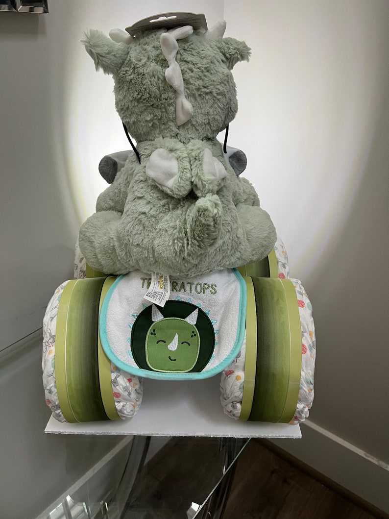 May include: A green plush dinosaur toy riding a diaper cake decorated with green and white stripes. The cake is labeled "TRICERATOPS" and has a green and blue bib with a dinosaur face on it.