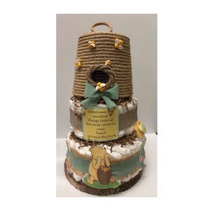 May include: A three-tiered diaper cake with a Winnie the Pooh theme. The top tier is a rope-wrapped beehive with decorative bees. The middle tier has a green ribbon and a sign that reads "Sometimes the smallest things take up the most room in your heart - Winnie the Pooh."