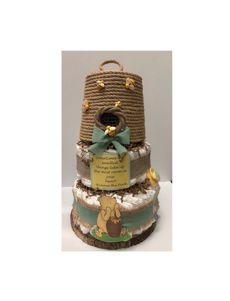 May include: A three-tiered diaper cake with a Winnie the Pooh theme. The top tier is a rope-wrapped beehive with decorative bees. The middle tier has a green ribbon and a sign that reads "Sometimes the smallest things take up the most room in your heart - Winnie the Pooh."