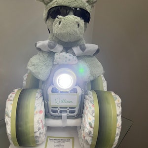 May include: A green stuffed animal wearing sunglasses sits on a diaper cake shaped like a four-wheeler. The cake is decorated with green and white fabric and has a white tag that says "William".