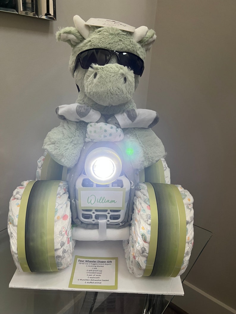 May include: A green stuffed animal wearing sunglasses sits on a diaper cake shaped like a four-wheeler. The cake is decorated with green and white fabric and has a white tag that says "William".