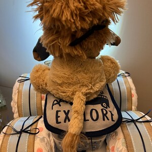 May include: A plush lion toy sitting in a diaper cake shaped like a car. The car is decorated with blue and white ribbon and the lion is wearing a bib that says "Go Explore".