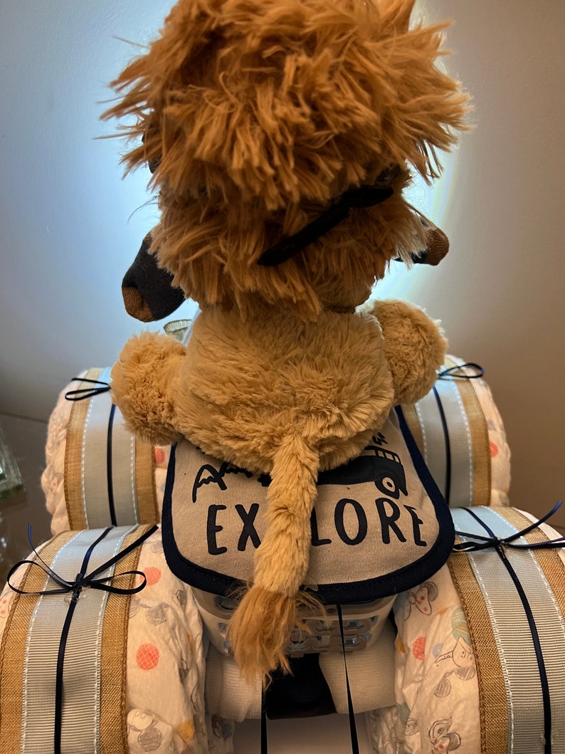May include: A plush lion toy sitting in a diaper cake shaped like a car. The car is decorated with blue and white ribbon and the lion is wearing a bib that says "Go Explore".