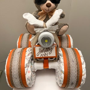 May include: A 4-wheeler diaper gift with a teddy bear wearing sunglasses. The gift includes 55 size 2 diapers, 2 washcloths, 1 bib, 1 receiving blanket, 1 teddy bear with sunglasses, and 1 Munchkin deluxe baby dishwasher basket. The gift is decorated with brown and orange ribbon and lace.