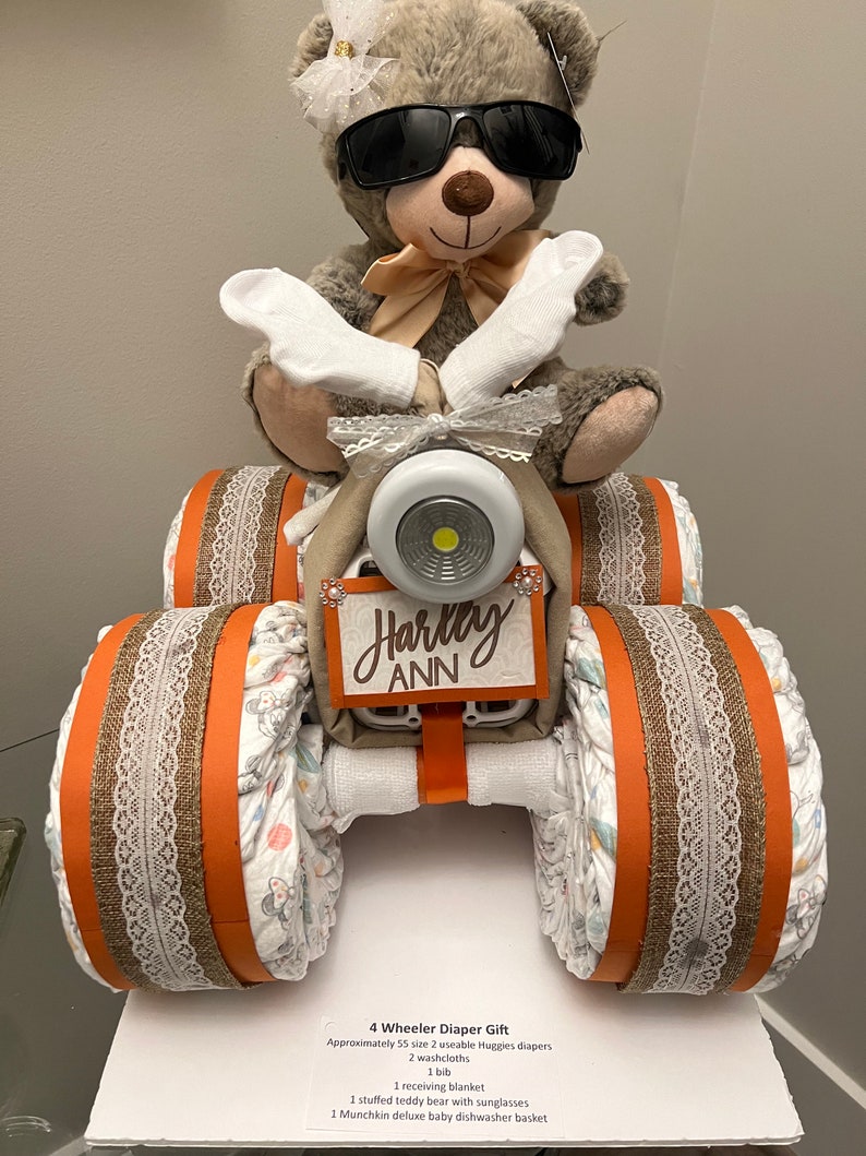 May include: A 4-wheeler diaper gift with a teddy bear wearing sunglasses. The gift includes 55 size 2 diapers, 2 washcloths, 1 bib, 1 receiving blanket, 1 teddy bear with sunglasses, and 1 Munchkin deluxe baby dishwasher basket. The gift is decorated with brown and orange ribbon and lace.
