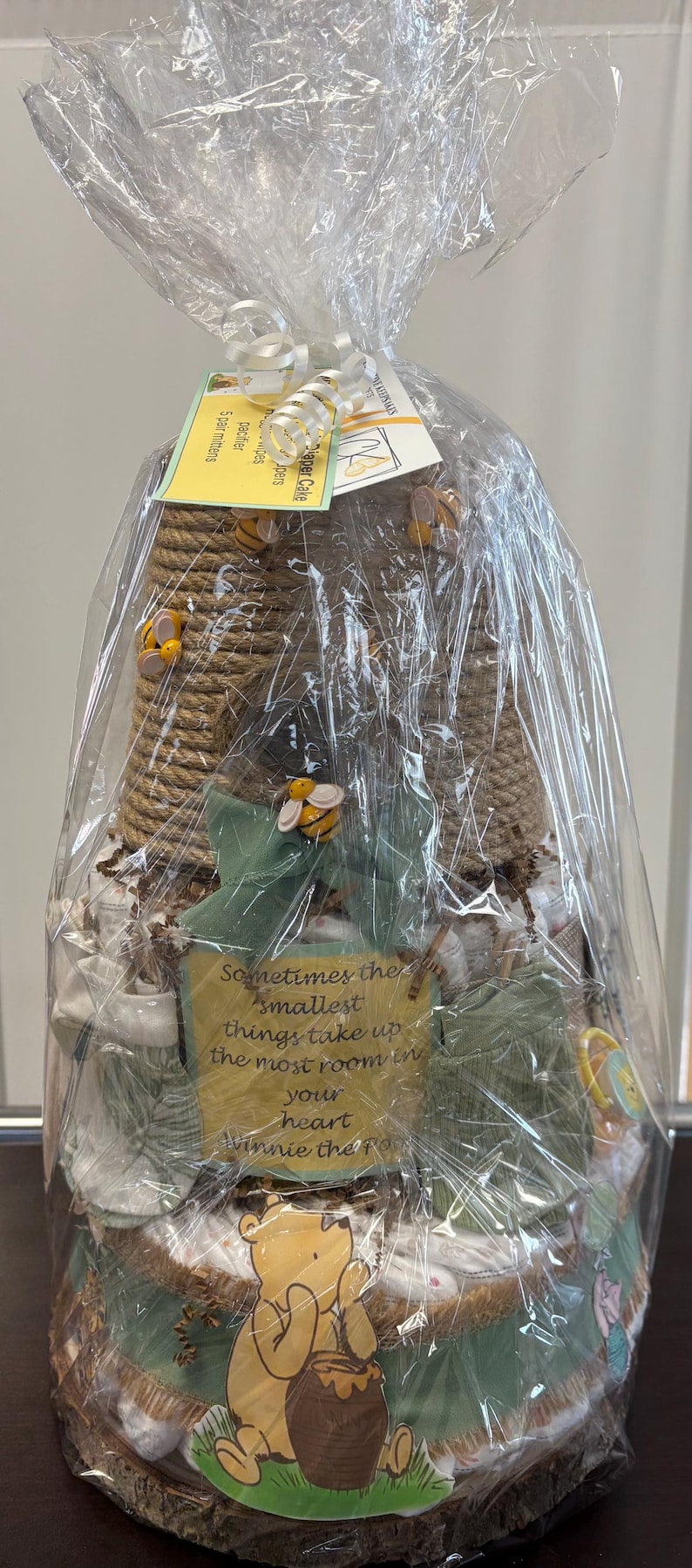May include: A Winnie the Pooh themed diaper cake, wrapped in clear cellophane. The cake features a beehive design, decorative bees, and a sign with a quote. The base has a Winnie the Pooh graphic.