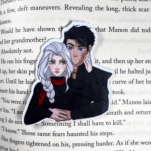 Puede incluir: Una pegatina que representa a una pareja con piel clara, cabello oscuro y ojos azules. La mujer tiene el pelo largo y blanco y una bufanda roja. El hombre la está abrazando. La pegatina está en una página de un libro.