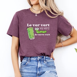 Puede incluir: Una camiseta morada con un gusano verde y un vaso de l&iacute;quido verde. El texto en la camiseta dice "Le ver vert va vers le verre vert".