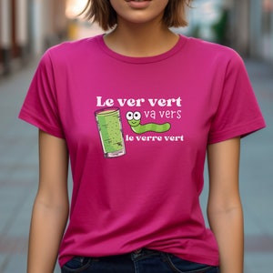 Puede incluir: Una camiseta rosa con un gr&aacute;fico blanco que dice "Le ver vert va vers le verre vert" y presenta un gusano verde con cara y un vaso de l&iacute;quido verde.