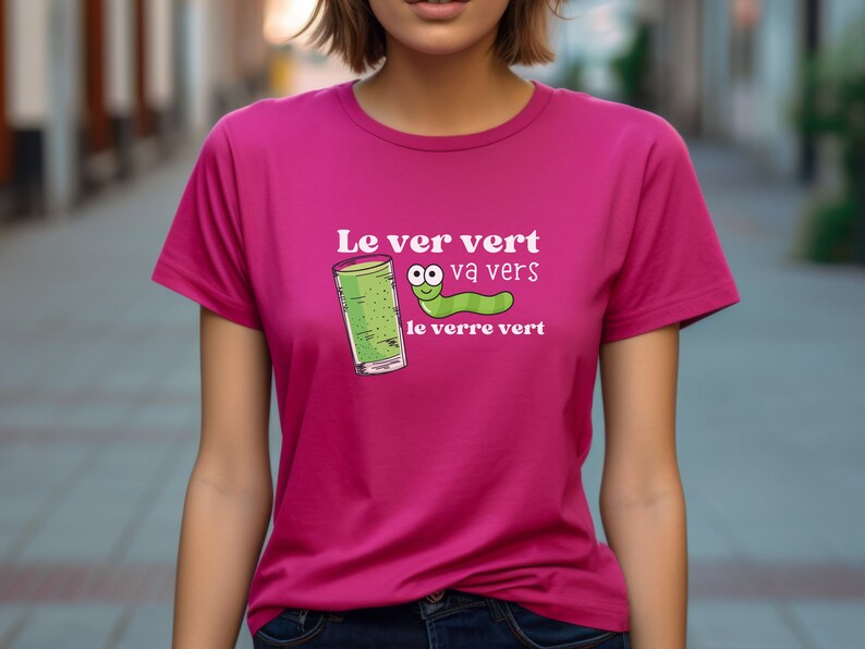 Puede incluir: Una camiseta rosa con un gr&aacute;fico blanco que dice "Le ver vert va vers le verre vert" y presenta un gusano verde con cara y un vaso de l&iacute;quido verde.