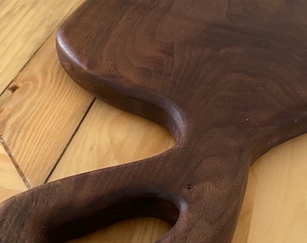 Puede incluir: Una tabla de cortar de madera grande, de color marrón oscuro, con un mango curvo único. La tabla está hecha de madera de nogal y tiene un acabado natural y rústico.
